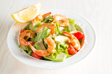 Fresh shrimp salad made of tomato, ruccola, avocado, prawns, chicken breast, arugula, crackers and spices. Caesar salad in a white, transparent bowl on wooden background