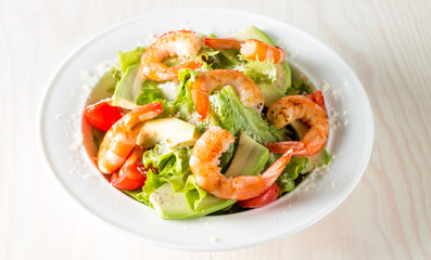 Fresh shrimp salad made of tomato, ruccola, avocado, prawns, chicken breast, arugula, crackers and spices. Caesar salad in a white, transparent bowl on wooden background
