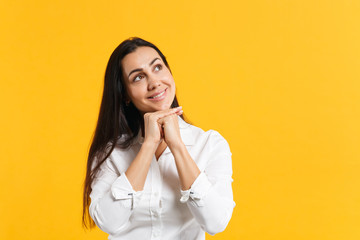 Portrait of pensive smiling young woman in white shirt put hand prop up on chin, looking up isolated on bright yellow orange wall background in studio. People lifestyle concept. Mock up copy space.