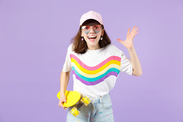Excited teen girl in vivid clothes, eyeglasses hold skateboard, keeping mouth open spreading hands isolated on violet pastel background. People sincere emotions, lifestyle concept. Mock up copy space.