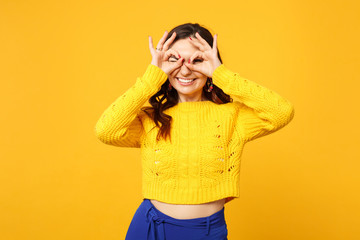 Funny blinking woman in sweater blue trousers hold hands near eyes imitating glasses or binoculars isolated on yellow orange background. People sincere emotions, lifestyle concept. Mock up copy space.