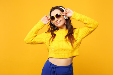 Attractive woman in sweater, blue trousers, sunglasses listen music with headphones looking aside isolated on yellow orange background. People sincere emotions, lifestyle concept. Mock up copy space.
