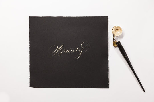 the word "Beauty" hand-written with silver ink, a tiny ink container and a nib mounted on a oblique holder are close to the paper.