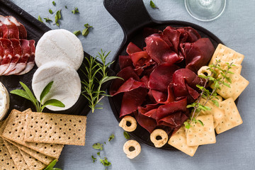 Italian bresaola served sliced on a tray on a table with white wine, crackers, grissini and taralli with aromatic herbs on a blue linen festive tablecloth.