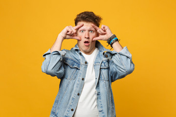Portrait of shocked young man in denim casual clothes keeping mouth open, stretching eyelids isolated on yellow orange wall background. People sincere emotions, lifestyle concept. Mock up copy space.