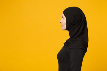 Side view image of beautiful young arabian muslim woman in hijab black clothes posing isolated on yellow wall background, studio portrait. People religious Islam lifestyle concept. Mock up copy space.
