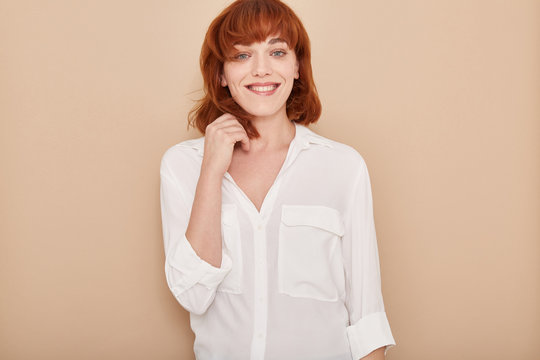Adult red haired and blue eyes woman portrait smiling anf touching her hair, dressing informal white T-shirt looking at camera over brown background