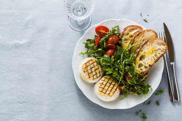 grilled Italian Tomino cheese served on a table with arugula salad and fresh homemade ciabatta bread and tomato on a blue linen festive tablecloth. summer menu
