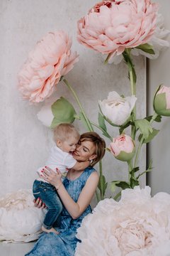 Charming mother hugging her little son among giant artificial flowers indoors