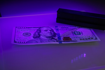 Verifying the authenticity of the hundred dollar bill in ultraviolet light. US banknotes, Benjamin Franklin. Equipment for in-depth inspection of money. Fake money or financial crisis concept