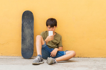 Front view of young boy sitting on ground leaning on a yellow wall while using a mobile phone