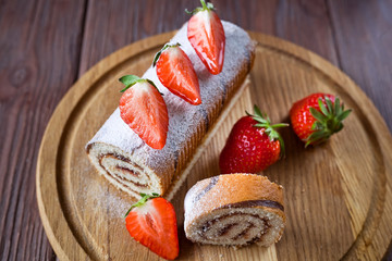 strawberry roll decorated with powder and fresh strawberries on the board, brown background, side view