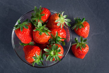 fresh tasty strawberries in a transparent bowl and two berries on a table on a dark gray background