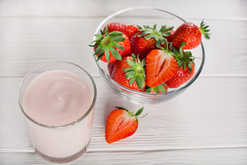 fresh strawberries in a transparent bowl and a glass of yogurt or cocktail on a white wooden table,...