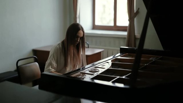 Young woman pianist playing gentle classical music on grand piano near the window. Talented girl pianist with long straight hair masterfully plays on the piano. Girl wears choker around her neck