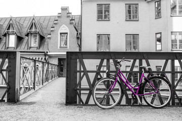 A picture of a lonely purple bike standing in the typical street in Stockholm by the bridge to a house. The bike looks to be modern in a retro style. The background is black and white. 