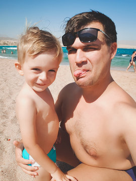 Funny image of young man with his little son showing tongues in camera while making selfie photo at sea beach. Family relaxing and having good time during summer holiday vacation.