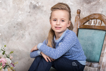 Portrait of a little attractive smiling girl in a blue sweater and pants with her hair folded in her hair sitting on a chair against a grunge wall background.