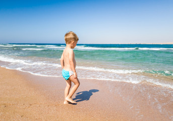 Cute little boy walking to the sea and looking at ocean waves. Child relaxing and having good time during summer holiday vacation.