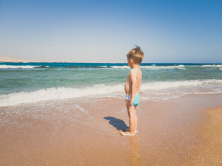 Fototapeta na wymiar Toned image of 3 years old toddler boy standing on the sea beach and looking at horizon. Child relaxing and having good time during summer holiday vacation.