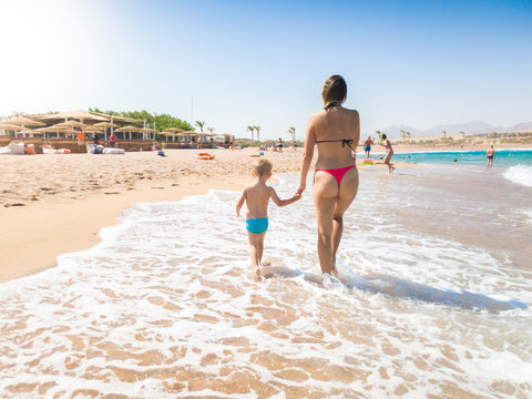 Image of happy young mother walking with her toddler son at the beautiful sea beach. Family relaxing and having good time during summer holiday vacation.