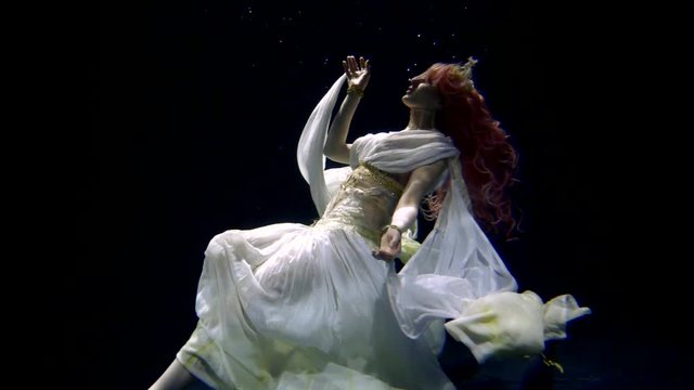 woman wearing in silk dress is keeping afloat inside water of sea in darkness, looking up
