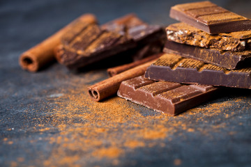 Chocolate stacked on black background. Chocolate bar pieces heap witn cinnamon powder and sticks. Sweet food photo concept.