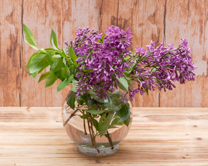 Fresh cut Purple Lilac Flowers in clear glass vase on wooden background. Syringa vulgaris.