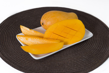Yellow mango and slices of mango are on the white plate. Brown tablecloth is under the plate