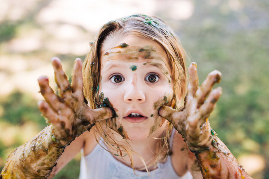 Toddler girl covered in paint making a funny face with her hands by her face.