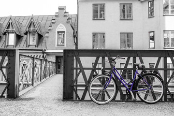 A picture of a lonely blue bike standing in the typical street in Stockholm by the bridge to a house. The bike looks to be modern in a retro style. The background is black and white. 