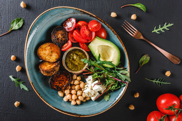 Fototapeta na wymiar Healthy Buddha bowl dish with avocado, tomato, cheese, chickpea, fresh arugula salad, baked potatoes and sauce pesto in black background. Dieting food, clean eating, top view.