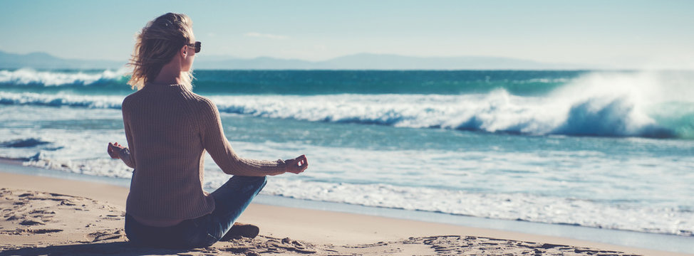 Horizontal image rear view young blond woman sitting in lotus position folded fingers mudra gesture meditating on the beach near the sea or ocean, water and clear sky copy space for ad concept text