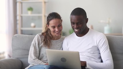 Happy mixed ethnicity young couple using laptop sitting on sofa