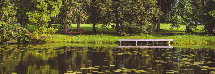 Horizontal image panoramic scenic view summer green landscape with picturesque forest lush lawn wooden pier dock for boats, in pond lake mirrored reflected nature, sunny day, Baltic States