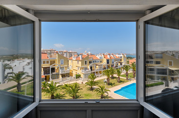 Through open window view to closed area of urbanization with planted palm trees on green lawn around swimming pool with blue water, yellow residential town houses with orange roofs, Torrevieja, Spain
