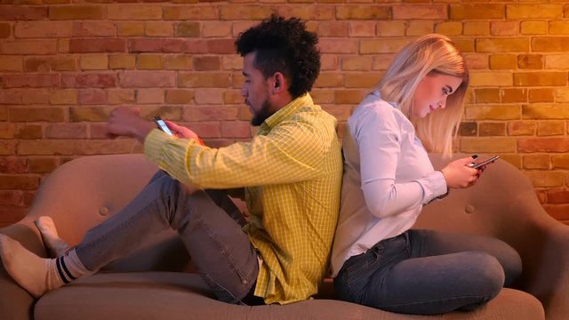 African guy and caucasian girl sitting back to back on sofa watching into smartphones at home.