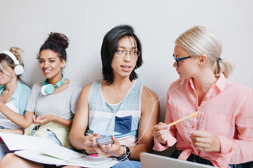 Asian guy with long hair discussing about new song with blonde female friend in glasses while black girl smiling on background. Indoor portrait of students enjoying music and joking.