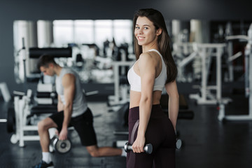 Fototapeta na wymiar Young sportswoman lifting weights in gym wearing sportswear with her boyfriend on background.