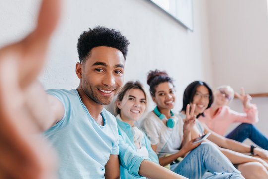 Glad black guy with stylish haircut making selfie while his smiling friends posing with peace sign. Cheerful students having fun in campus after lecture and laughing.