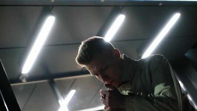 serious cool young man illuminated by white neon lamps smoking cigarette near the mall at night