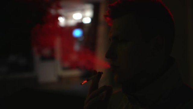 pensive young man smoking cigarette in the darkness illuminated by red neon lamp from behind on the street at night