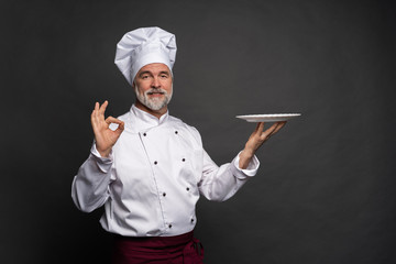 Mature cook chef holding an empty plate on a black background.