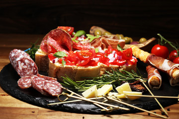 Traditional italian antipasto bruschetta appetizer with cherry tomatoes, cream cheese, basil leaves and balsamic vinegar on cutting board with prosciutto, salami, cheese