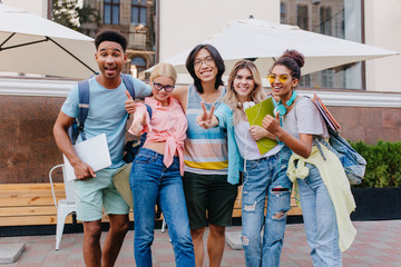 Fototapeta premium Happy blonde girl wears jeans with holes posing outdoor near smiling friends. Outdoor portrait of pleased students holding laptop and backpacks in morning.