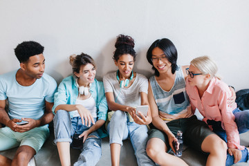 Pretty african girl with blue earphones showing her university mates something interesting in smartphone. International students sitting on floor and listening with interest what their friend tell.