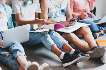 Girl in blue jeans and black sneakers writing lecture in big textbook, sitting on the floor with college friends. Young man typing message on phone while other students working with laptops.