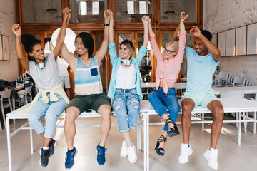 Fototapeta premium Joyful girl in jeans and black sneakers holding her asian friend's hand and smiling. Indoor portrait of happy students sitting on table together aftet work in library.