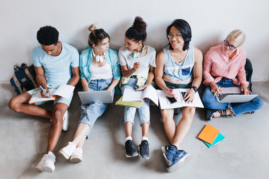 Overhead view of students of different nationalities sitting on the floor with laptops and notebook. Portrait of friends resting on the ground after examinations and talking.