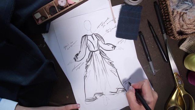 Top view of seamstress's hand working on fashion dress sketch in design workshop studio. Female tailor drawing sketch of dress ordered by customer in sewing atelier